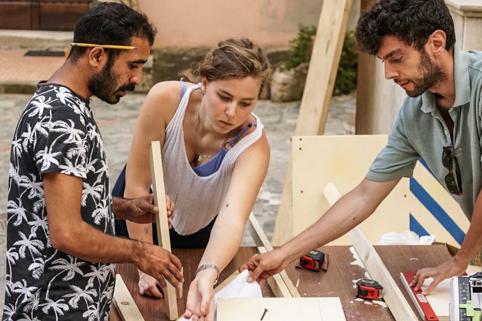 Tre persone assemblano listelli di legno in un cantiere allestito in strada, con avvitatore e livella sul banco da lavoro