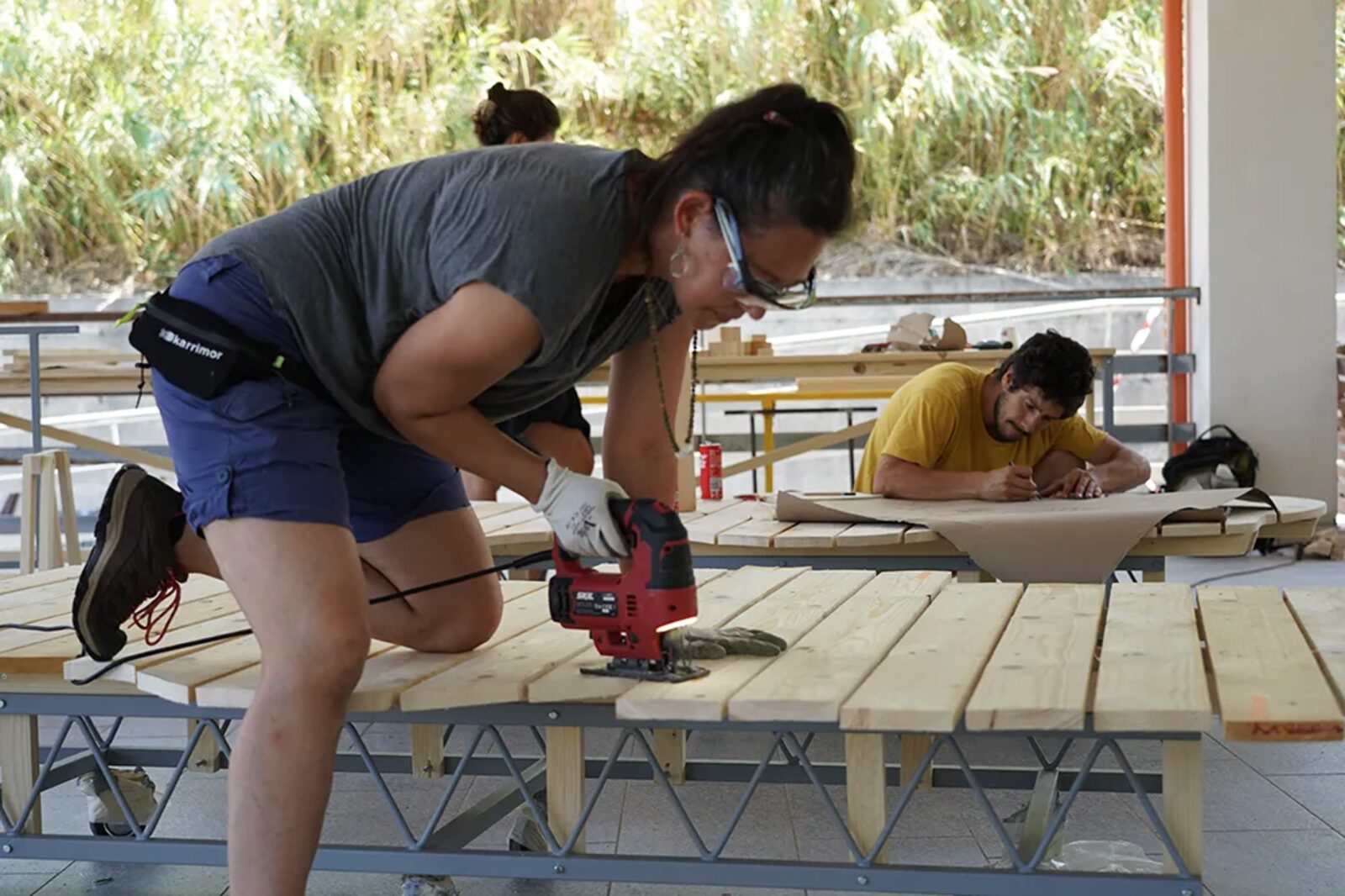 Una persona taglia col seghetto le tavole di legno di una seduta in costruzione, mentre altre due lavorano al disegno poco pi&ugrave; in l&agrave;