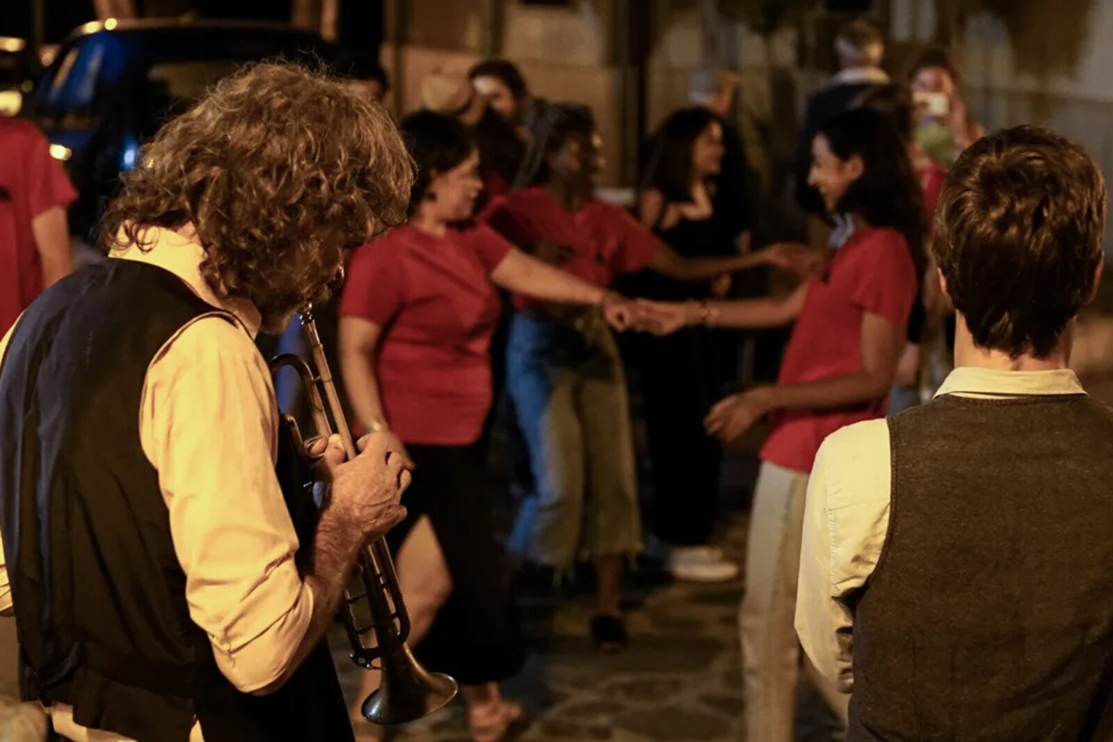 Una serata di festa all'aperto: un trombettista suona in primo piano mentre alle sue spalle alcune persone ballano