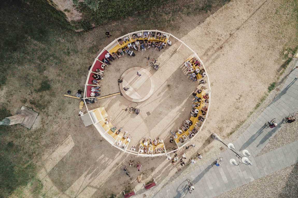 Veduta aerea di un grande tavolo circolare allestito in uno spazio pubblico, con molte persone sedute attorno